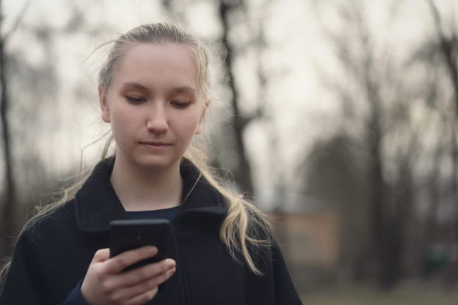 teen girl standing on sidewalk using smartphone in cold spring morning, copy space